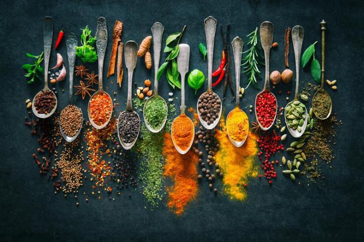 A beautiful array of rustic spoons are aligned at their rounded heads. They are resting on a black table, and the picture is taken from a birds eye view. There are spices of many different colors sprinkled artistically around. The light is shining on the silver spoons from directly above them.