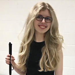 A photo of Kaleigh, a light-skinned woman with blonde hair and glasses, smiling at the camera. She wears a black dress, grips her cane in her right hand, and stands against a white backdrop.